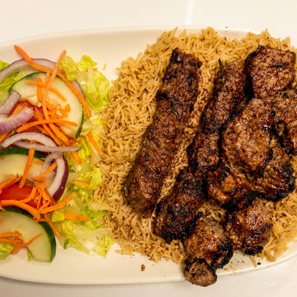 Grilled meat kebabs and chunks with pilaf rice and a fresh side salad on a white plate.
