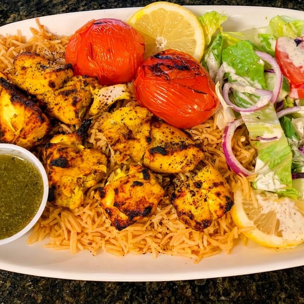 Grilled chicken, rice, roasted tomatoes, fresh salad, green sauce, and lemon slices on a white plate.
